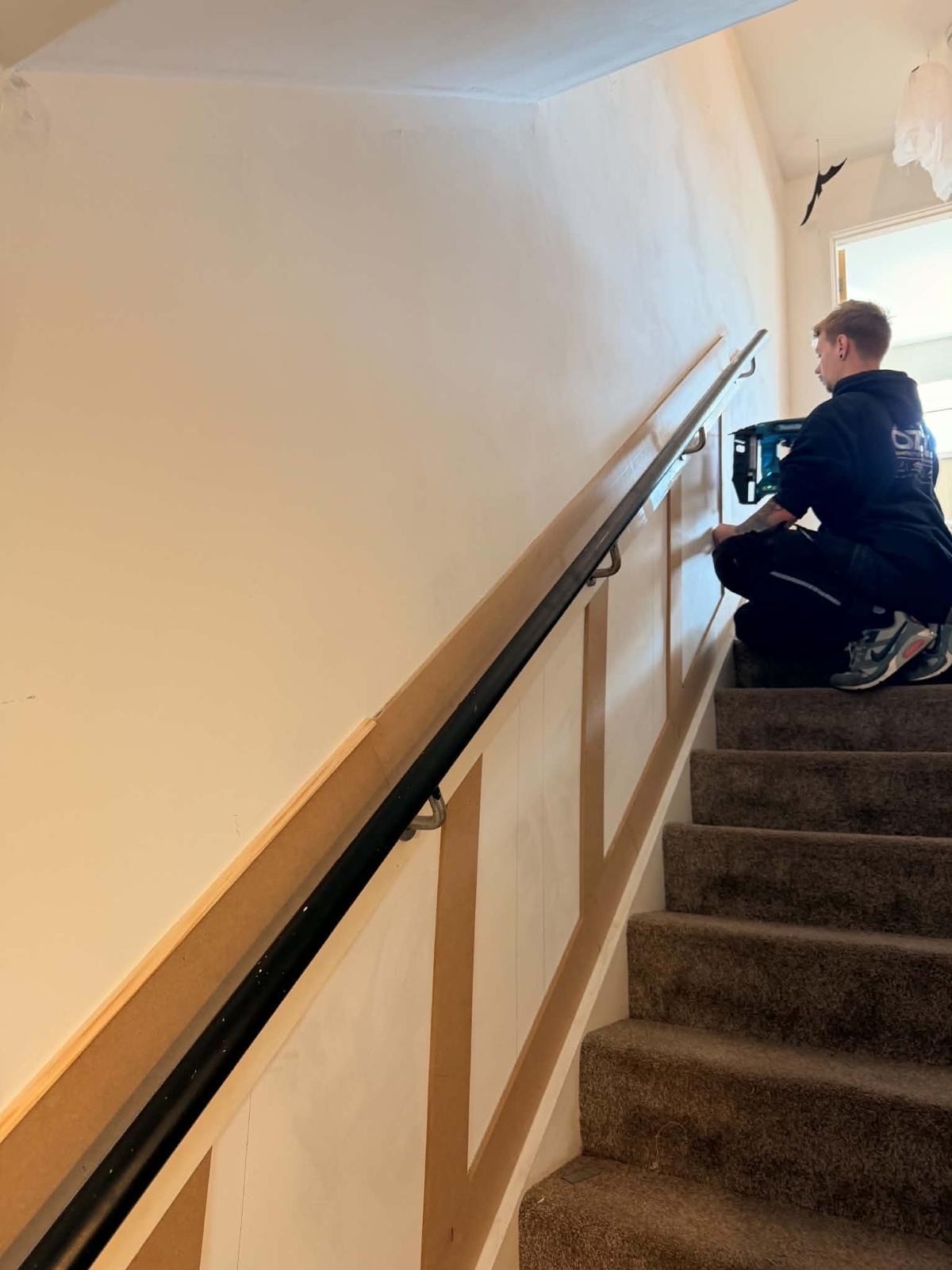 Ozz kneeling on a carpeted staircase, fixing angled timber panelling to the wall beneath the handrail with a cordless nail gun