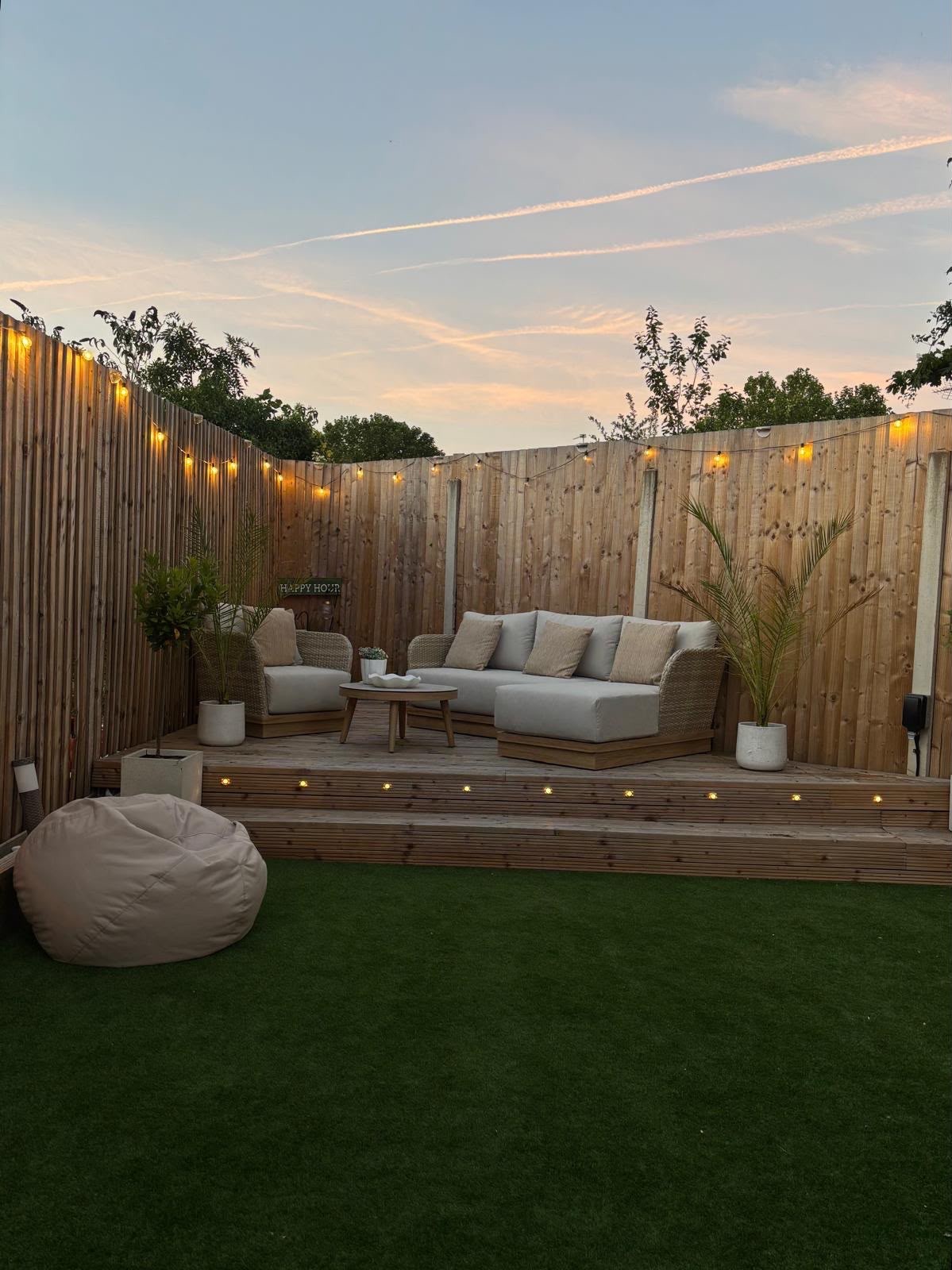 The same garden after the work: tiered timber deck with lit steps, artificial lawn, new fencing, festoon lights and an outdoor lounge