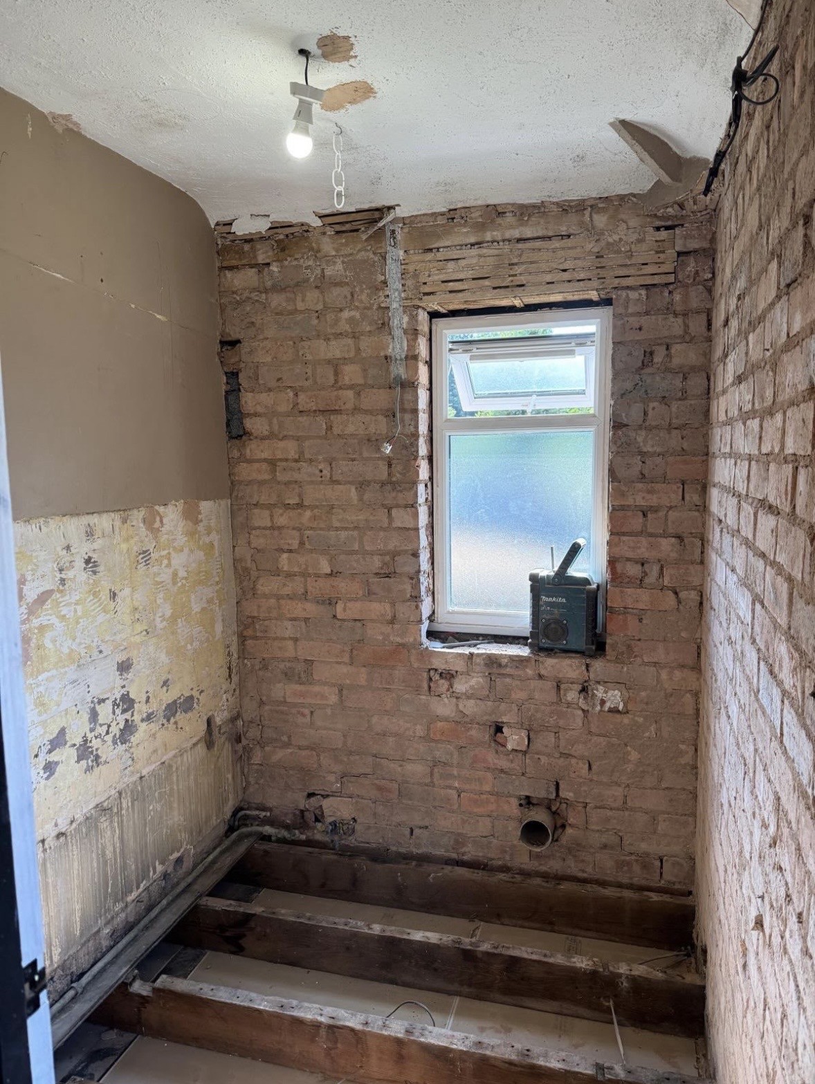 Bathroom before the work: stripped back to bare brick walls and exposed floor joists, with copper pipework and a single bare bulb