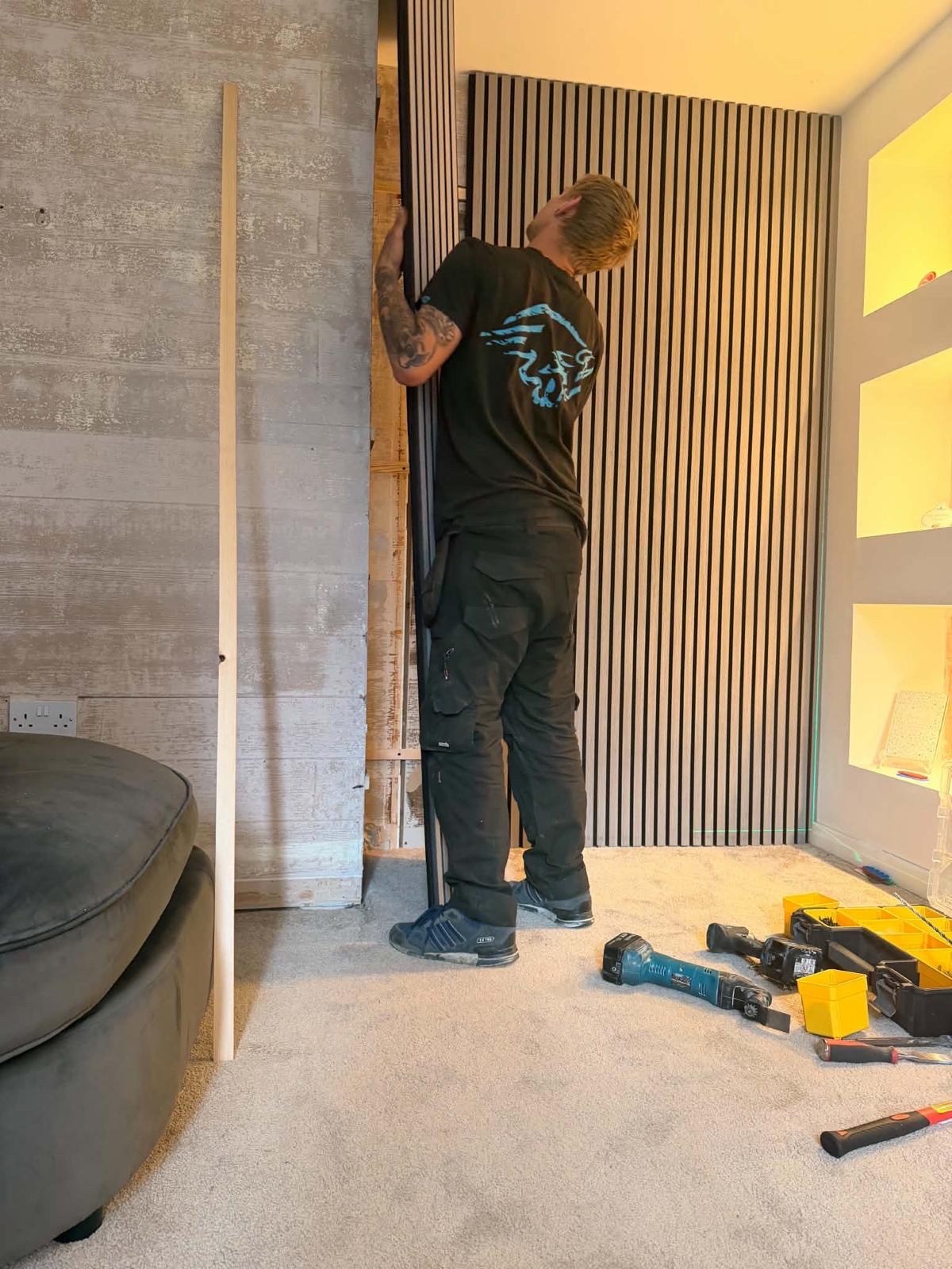 Ozz lifting a full-height acoustic slat panel into a lit alcove, with a green laser line marking the level